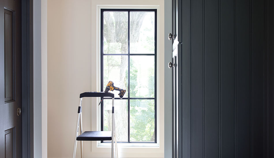 A stepladder with a drill resting in a holder stands in front of a tall narrow window with a black frame