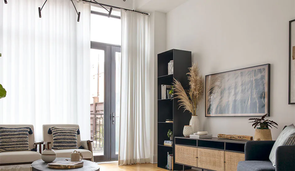 A living room has tall glass doors with minimalist window treatments including Ripple Fold Drapery in Basket Sheer, Winter
