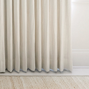 A panel of drapery made of Luxe Linen in Optic White hovers just above a white floor with a woven rug