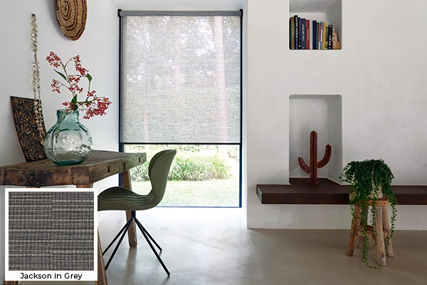 A rustic office with a unfinished wood desk has Roller Shades for windows made of Jackson in Grey over a tall window