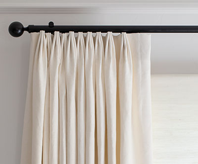 A close-up shot of oyster-colored thermal curtains, specifically tailored pleat drapery, hanging along a black rod