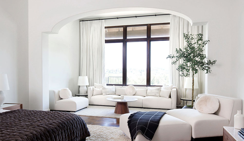 An elevated bedroom with a living area features blackout Tailored Pleat Drapery made of Linen in Ivory over a tall window