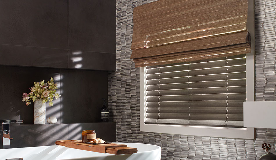 A bathroom with dark tiles has woven shades made of Artisan Weaves Del Rey in Taupe layered over Wood Blinds